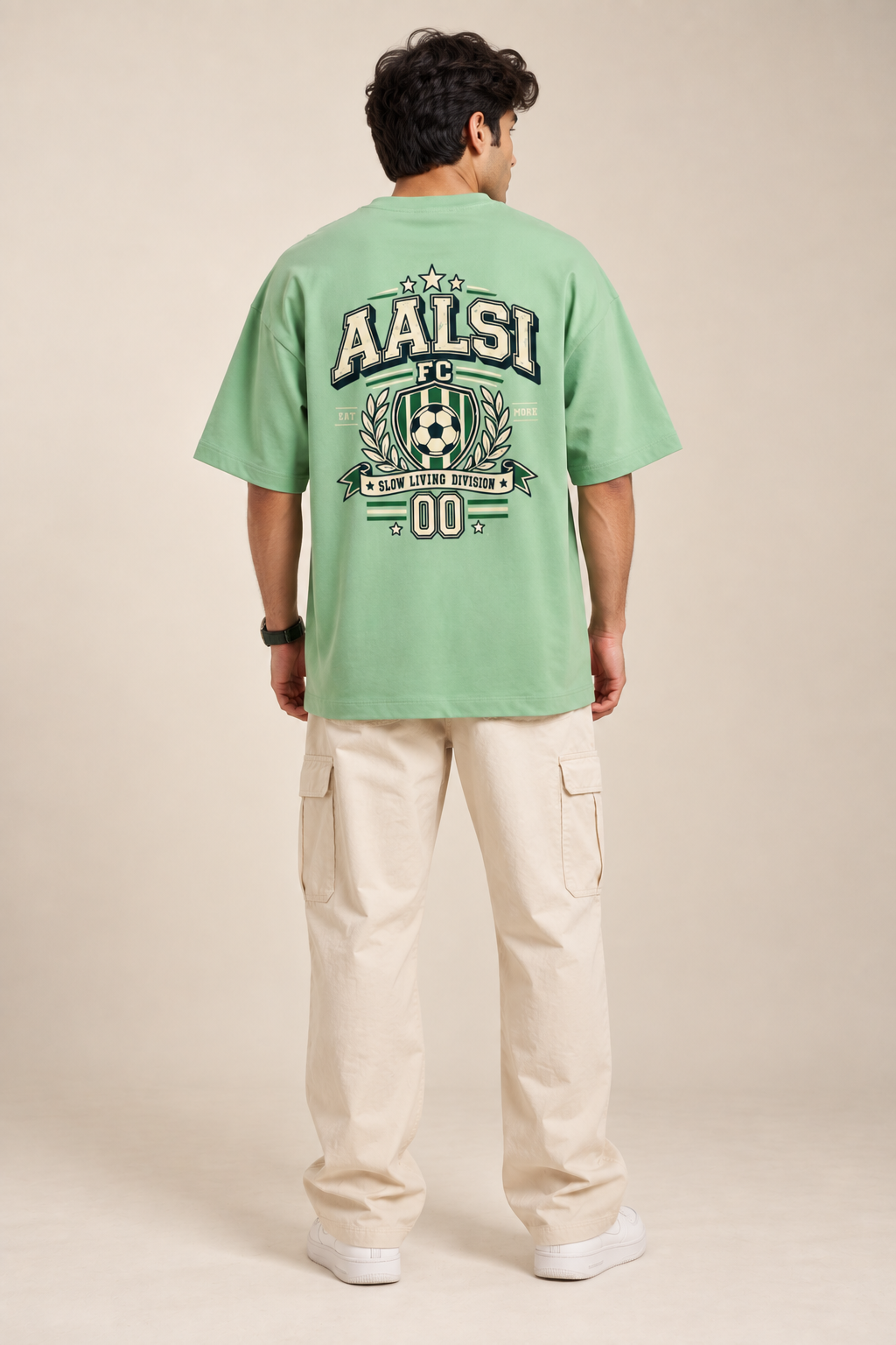 Jade green Aalsi FC oversized T-shirt featuring a varsity football crest, soccer ball, laurel graphics, and Slow Living Division slogan on back styled with baggy pants.