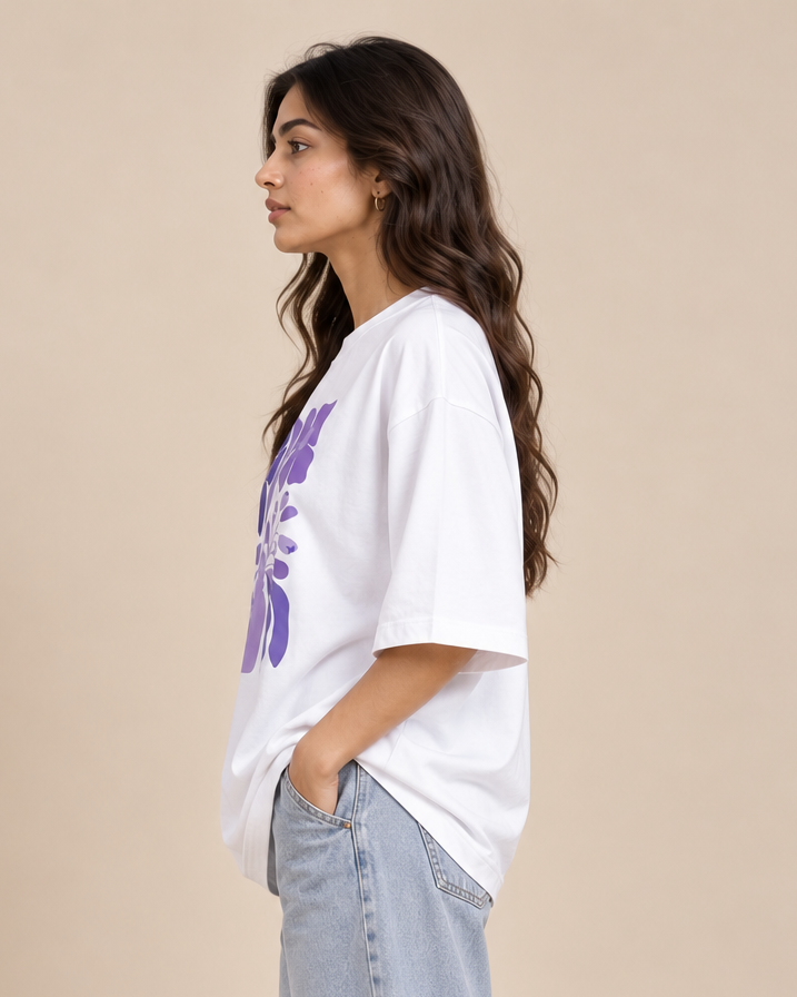 A woman in profile wearing an oversized white t-shirt with a purple floral graphic on the front, paired with light-wash jeans, set against a solid beige background.