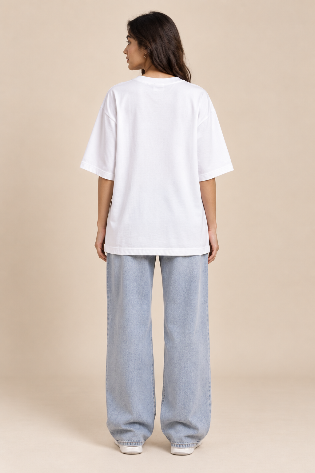 A woman is shown from the back wearing an oversized white short-sleeve t-shirt and light-wash wide-leg jeans, standing against a solid beige studio background.
