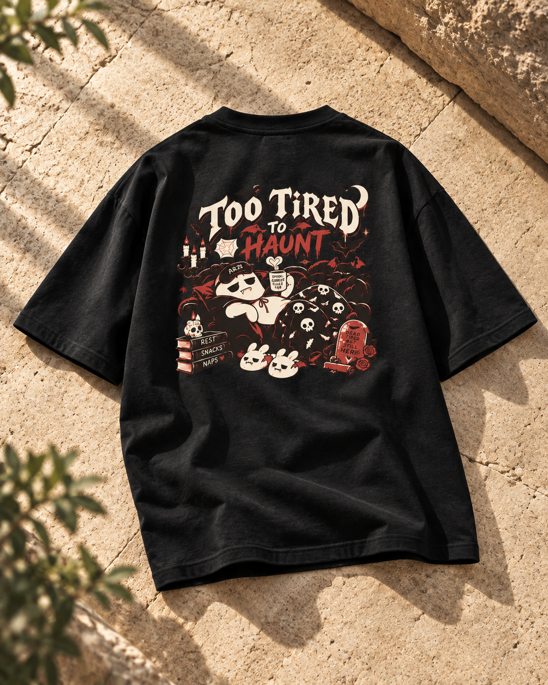 Back of a black oversized Halloween T-shirt featuring “Too Tired to Haunt” lettering, sleepy ghost characters, bats, skulls, candles, and red-and-cream gothic artwork.