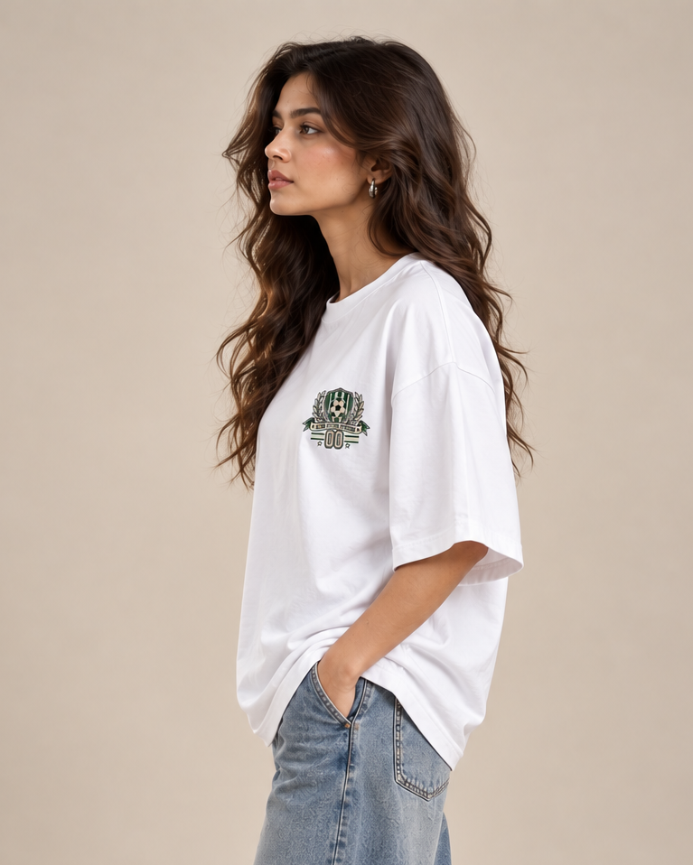 White oversized football T-shirt with a small green and cream soccer crest graphic on the chest styled with baggy pants.