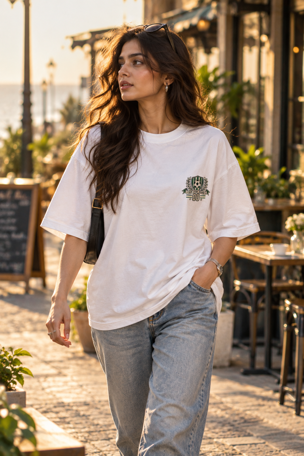 White oversized football T-shirt with a small green and cream soccer crest graphic on the chest styled with baggy pants.