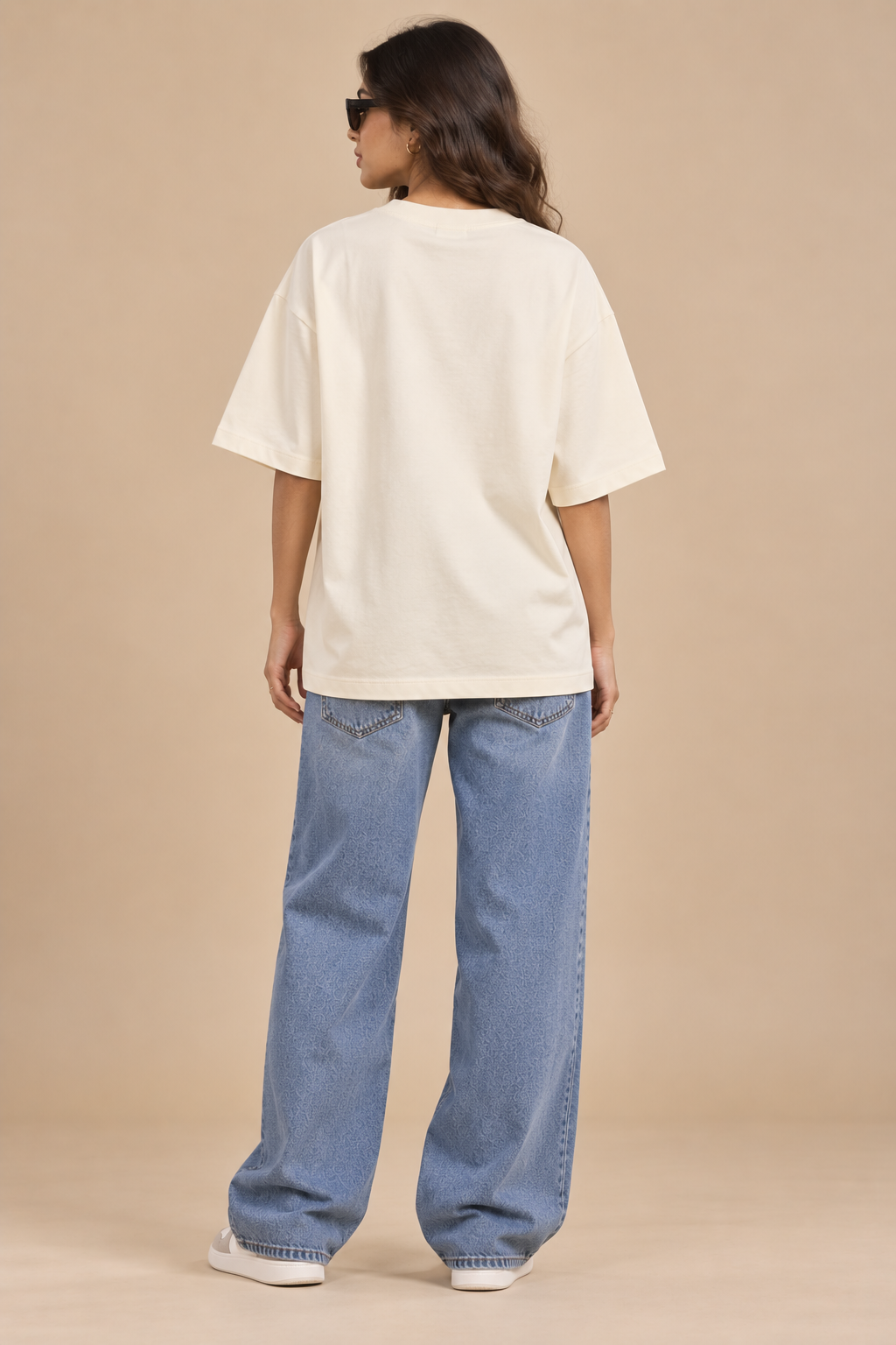 A woman shown from the back wearing an oversized cream-colored t-shirt, wide-leg light blue jeans, and white sneakers, standing against a solid beige studio background.