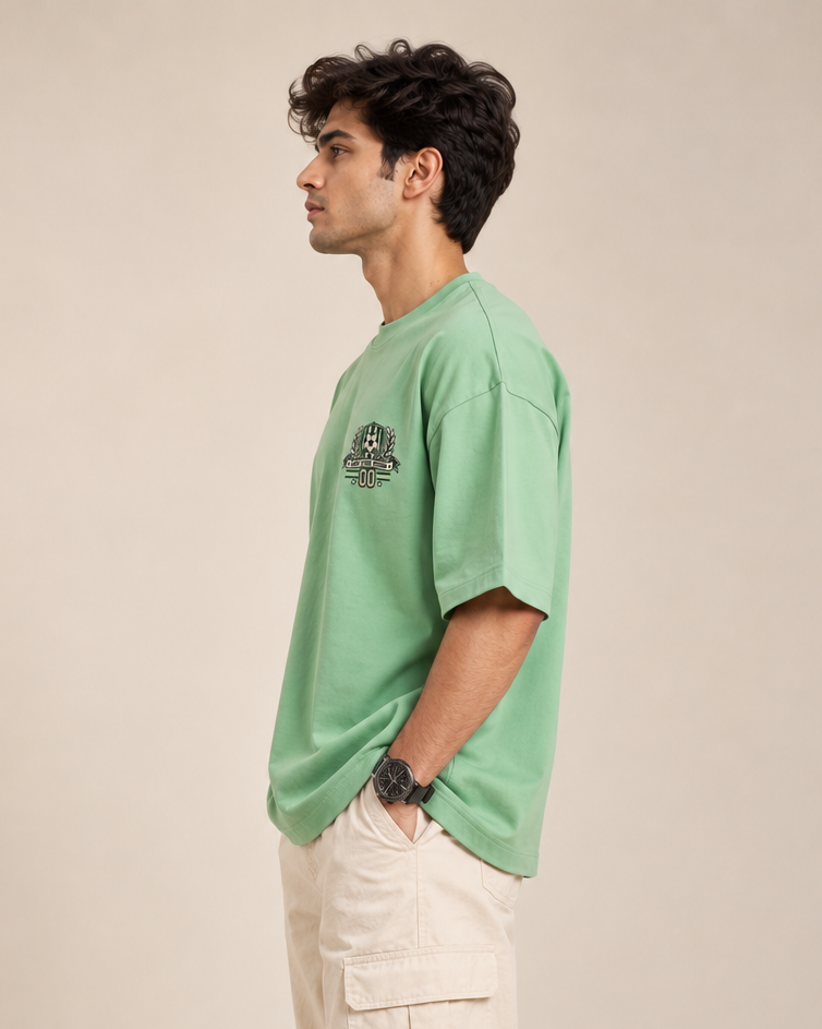 Jade green oversized football T-shirt with a small green and cream soccer crest graphic on the chest styled with baggy pants.
